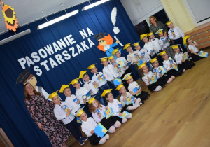 Dzieci w biretach na głowie z dyplomikami w rękach i uśmiechem na twarzy wraz z Paniami pozują do wspólnej fotografii